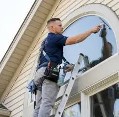 Professional window cleaner on a ladder using a squeegee to clean an exterior arched window, Ann Arbor, MI.